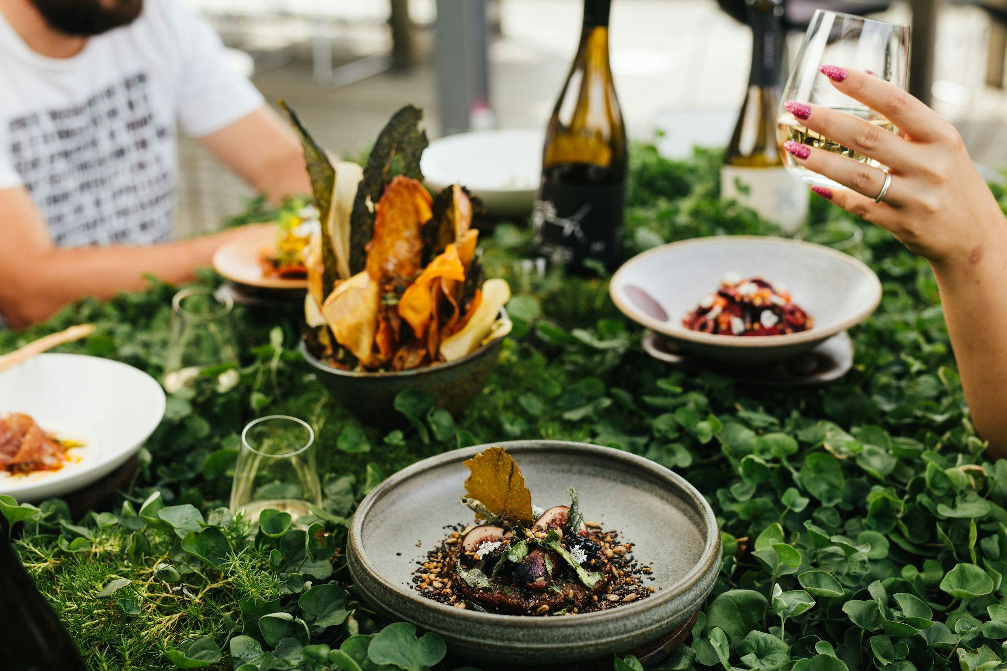 People dining on a lush tabletop of leaves with ceramic plates containing vibrant thoughtfully plated food.