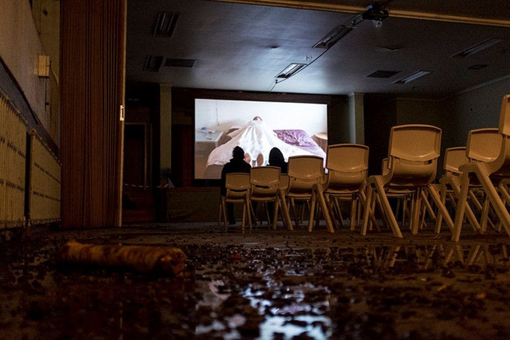 Two people watching a video of a person under a sheet on a bed. They are sitting in a dark, dirty room with rows of chairs directed towards the screen.