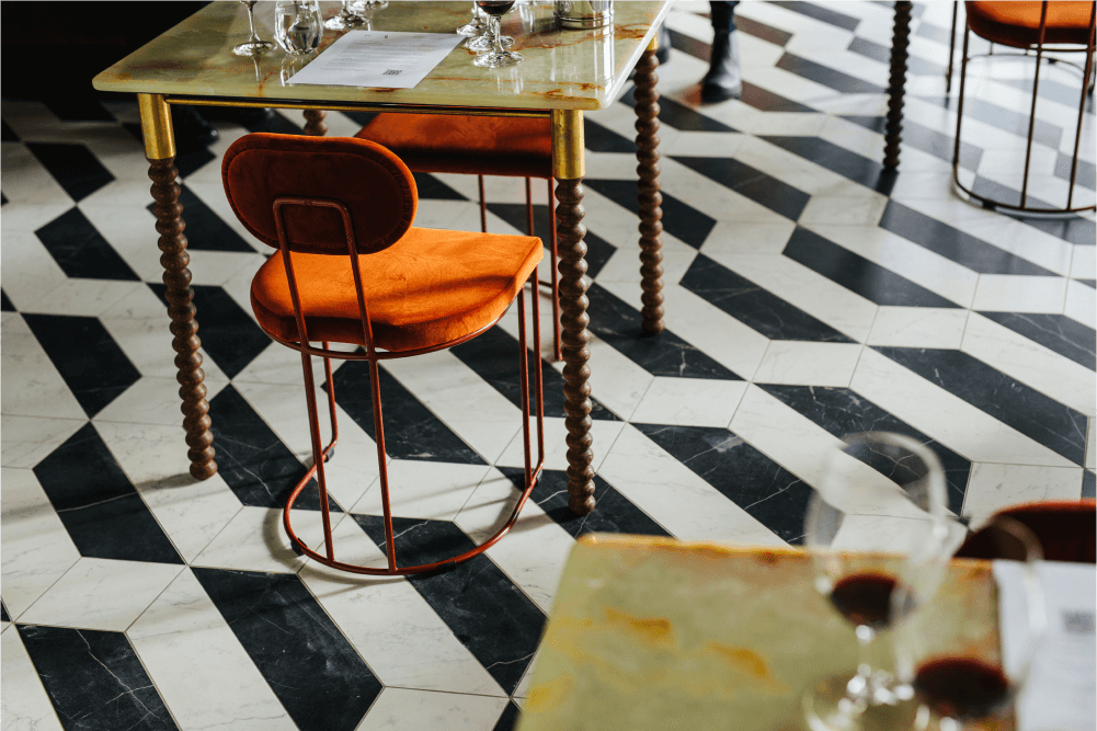 A black and white tiled floor, marbled table tops with ornate wooden legs and orange velvet seating