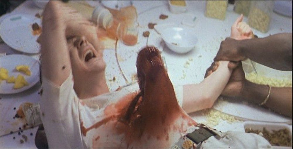Person lies on a food strewn table, held down while a toothed alien explodes from their stomach covered in blood.