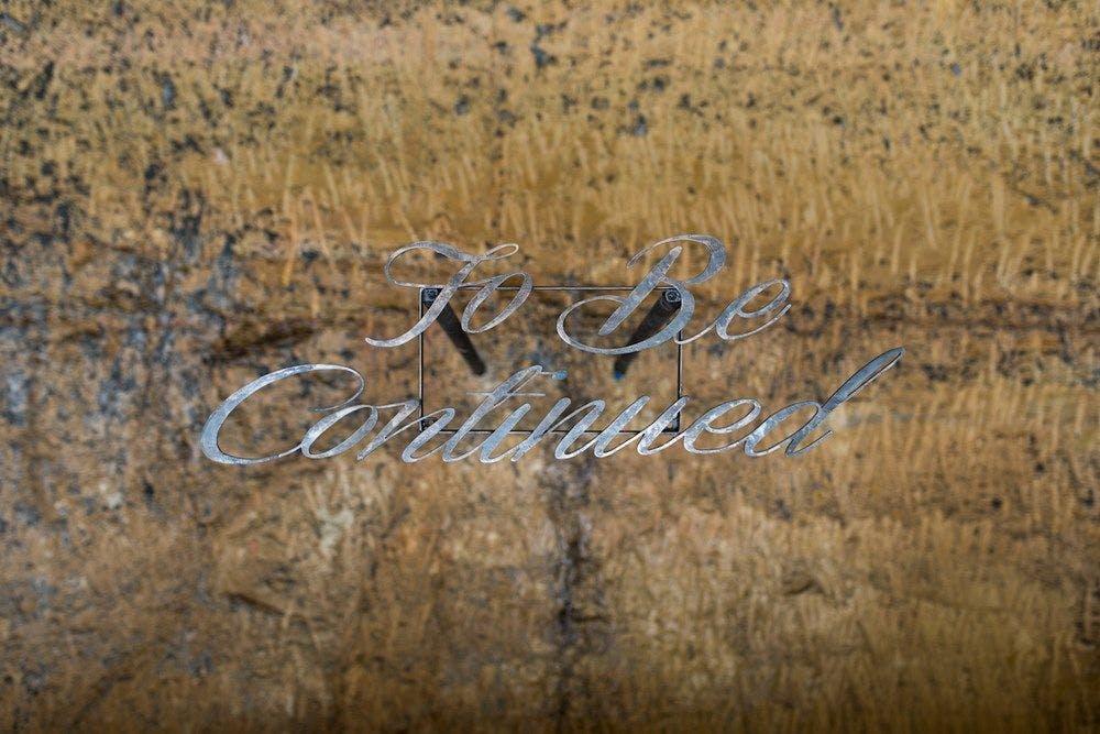 Metal sign with To Be Continued in cursive writing against a sandstone wall.