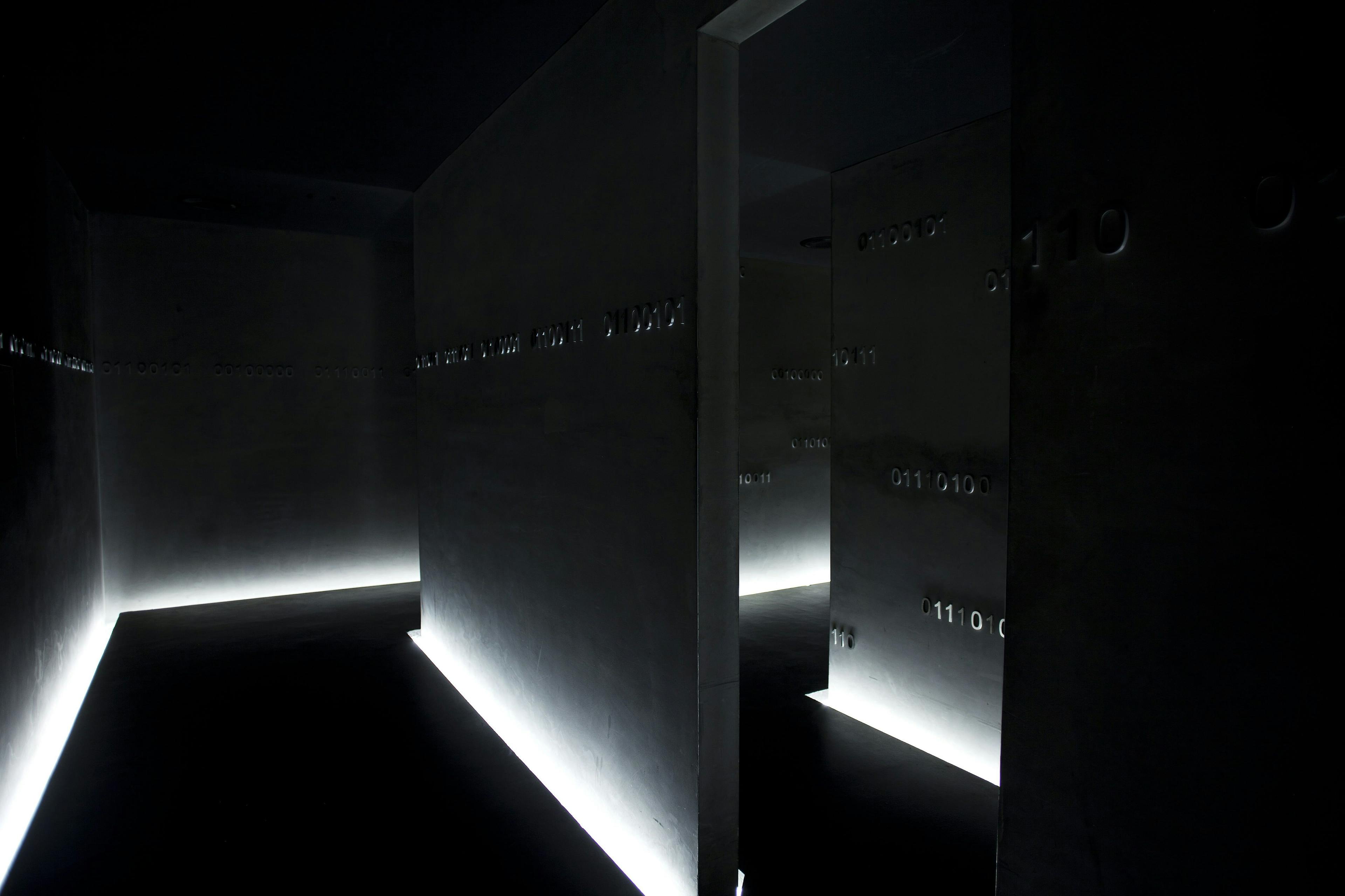 A dark passageway of thick grey walls lit from the bottom with binary code running along them.
