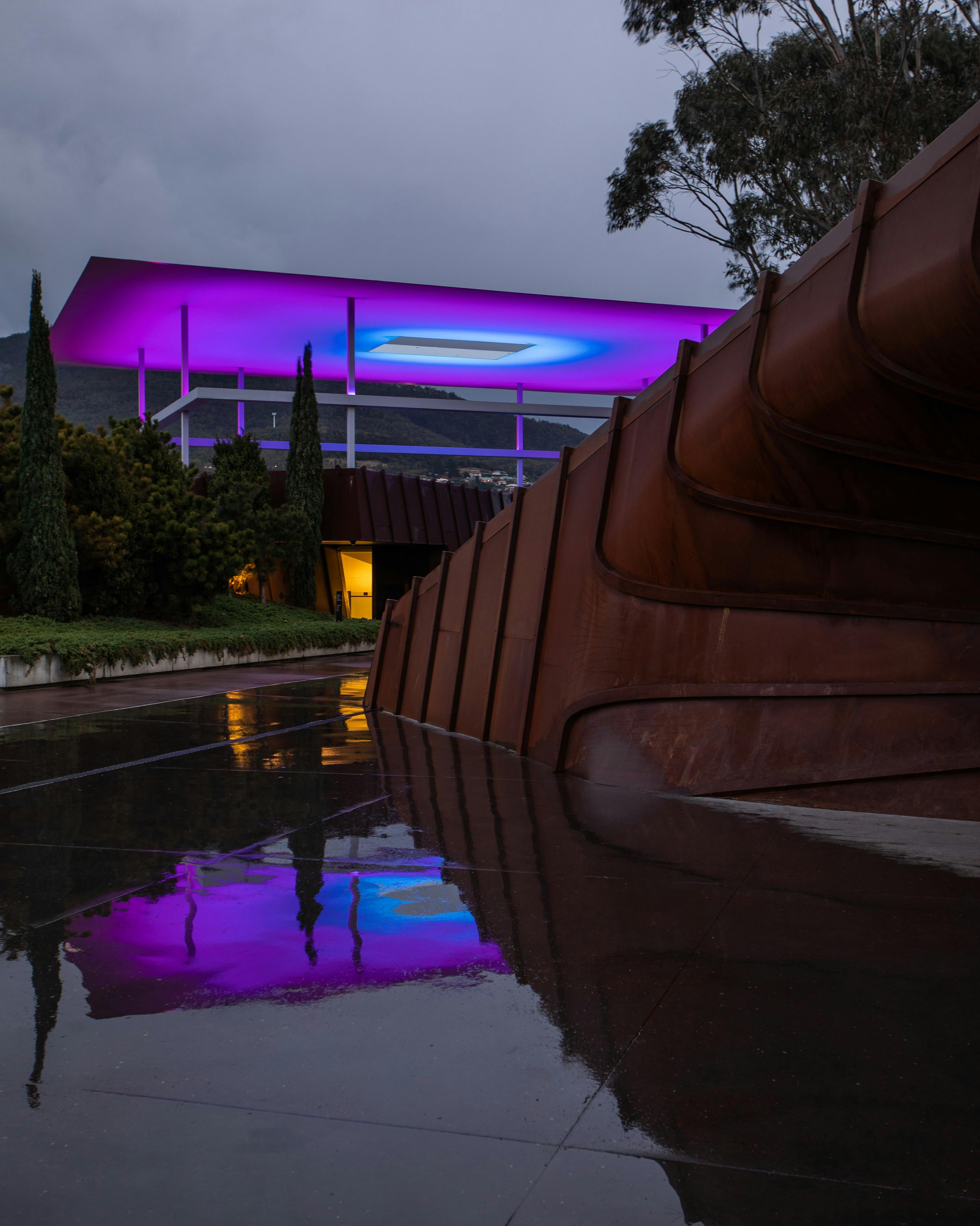 A large structure in the background shines hues of pinks and blues, reflecting off a puddle in front of a corten steel structure.
