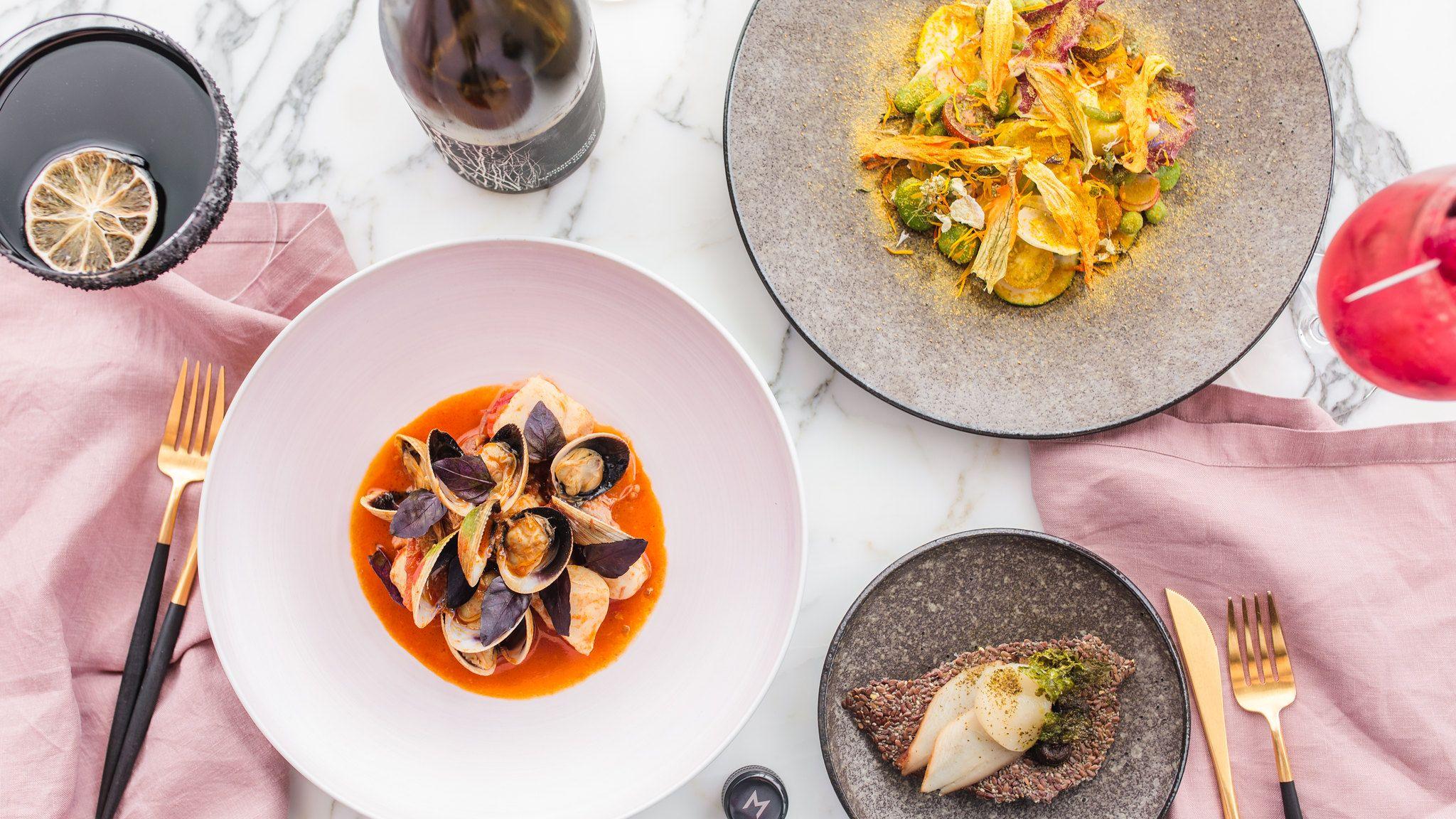 Birds eye view of a marble table set with a vibrant dish of clams, dark cocktails and gold cutlery