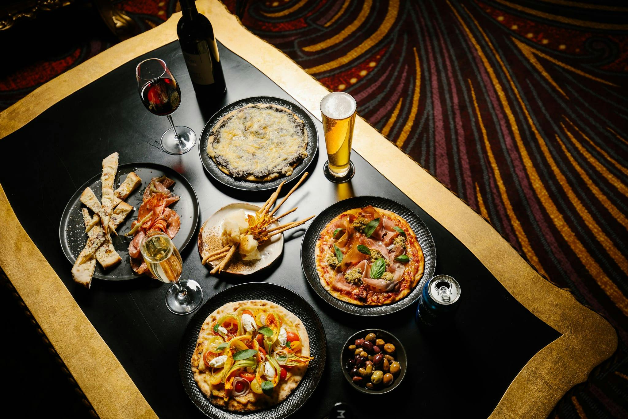 A black table with gold trim on luxurious carpet set with pizza, grissini, olives, wine and beer.