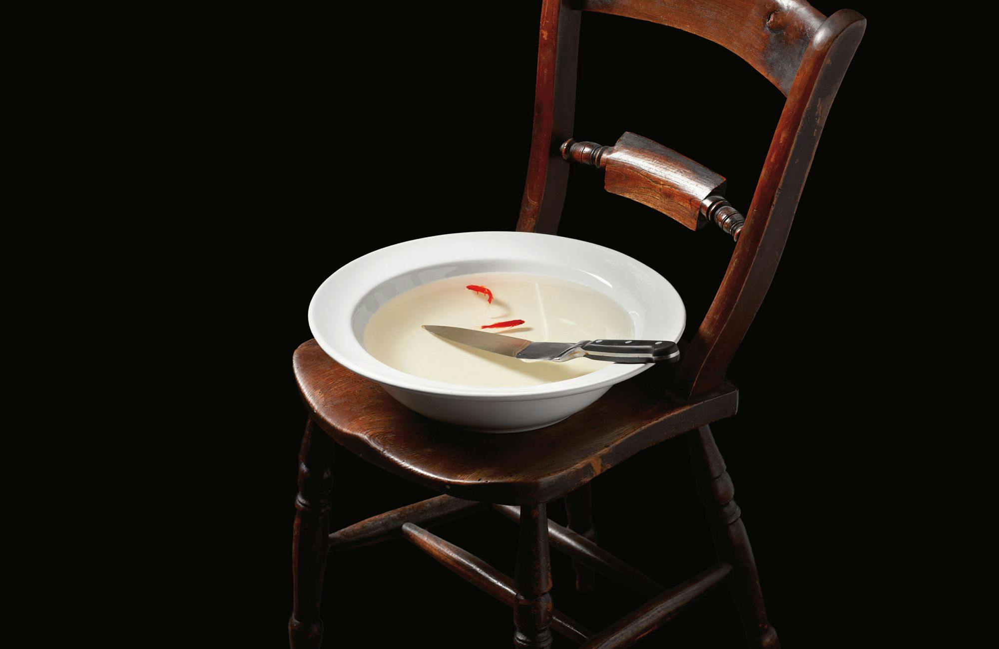 On a wooden chair sits a white bowl containing water, a knife and two gold fish.