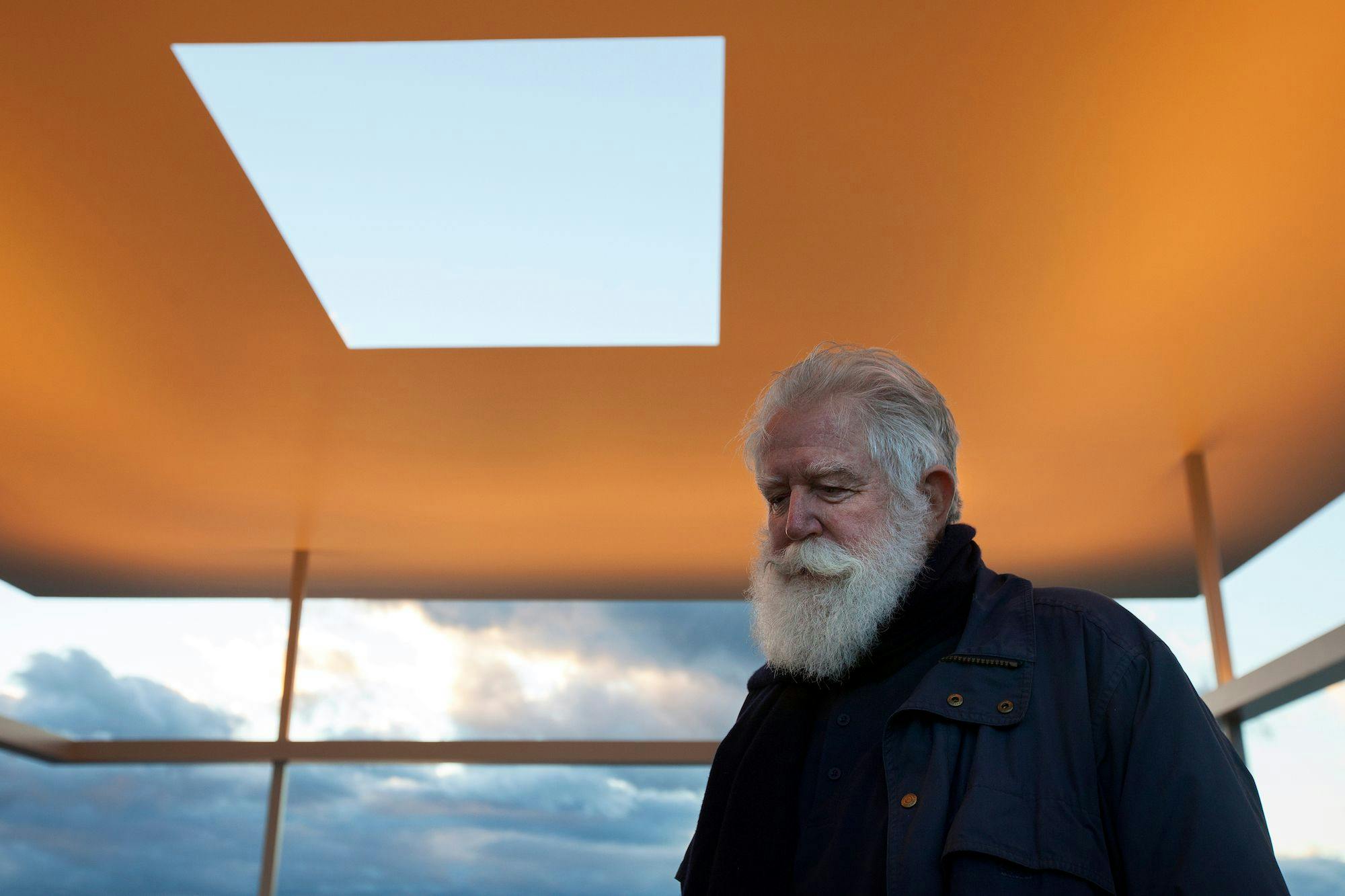 A white bearded James Turrell is gazing down at the ground. Above him, there is a large square hole in the roof, revealing the clouds in the sky beyond.