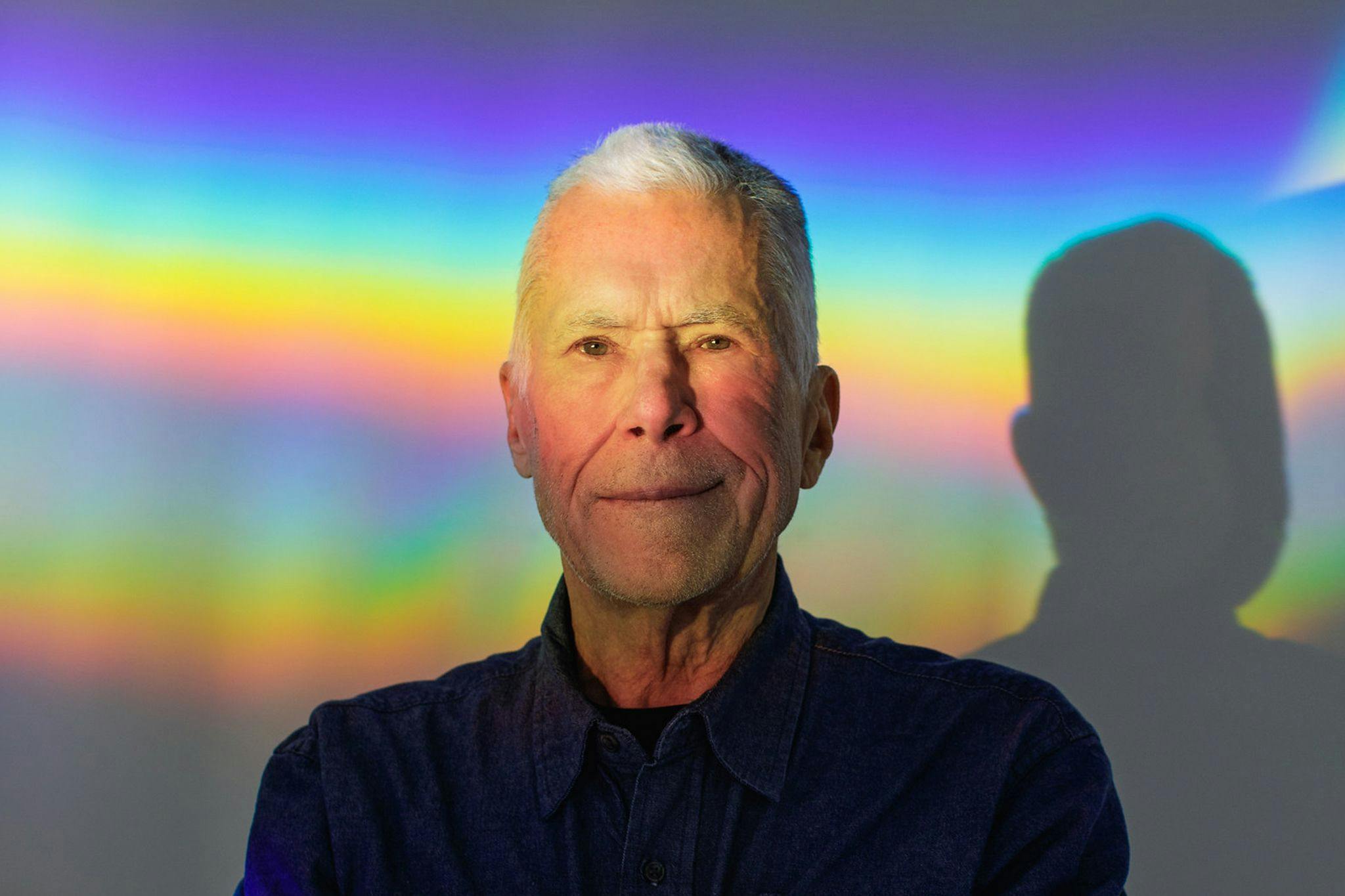 Charles Ross stands against a blank wall, with a spectrum of light casting over his face and the wall behind him.