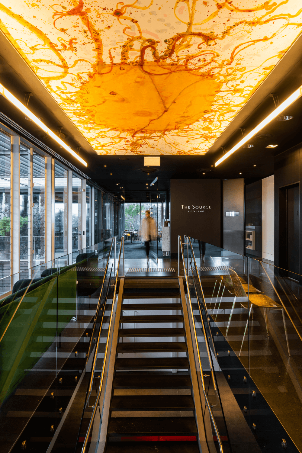 The entrance of the Source restaurant, featuring a staircase and a large abstract painting of the sun, illuminated from above.