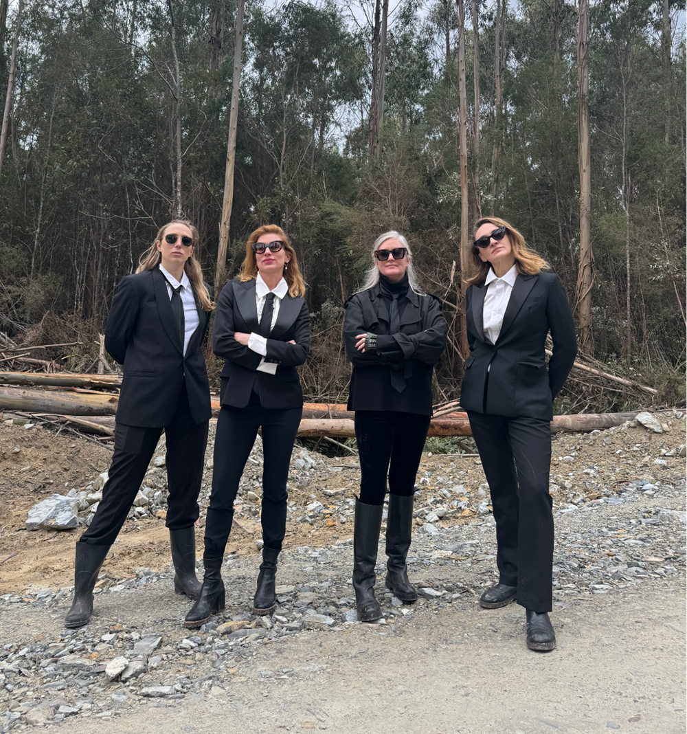 Four women dressed in black suits and sunglasses on a dirt road in front of trees.