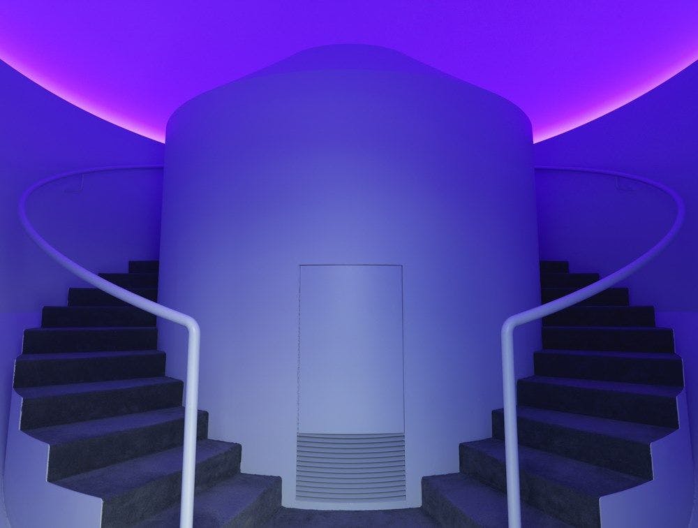 Two circular stair cases lead behind a white centre. Ceiling is lit with blue and pink hues.