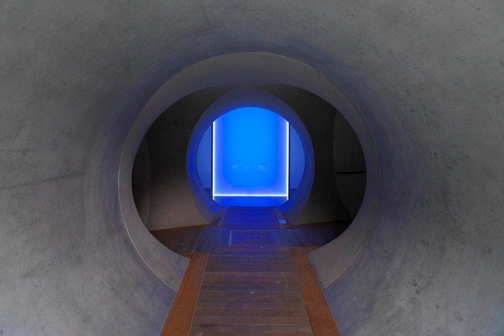A tunnel of concrete and wooden floor with a bright blue light at the end.
