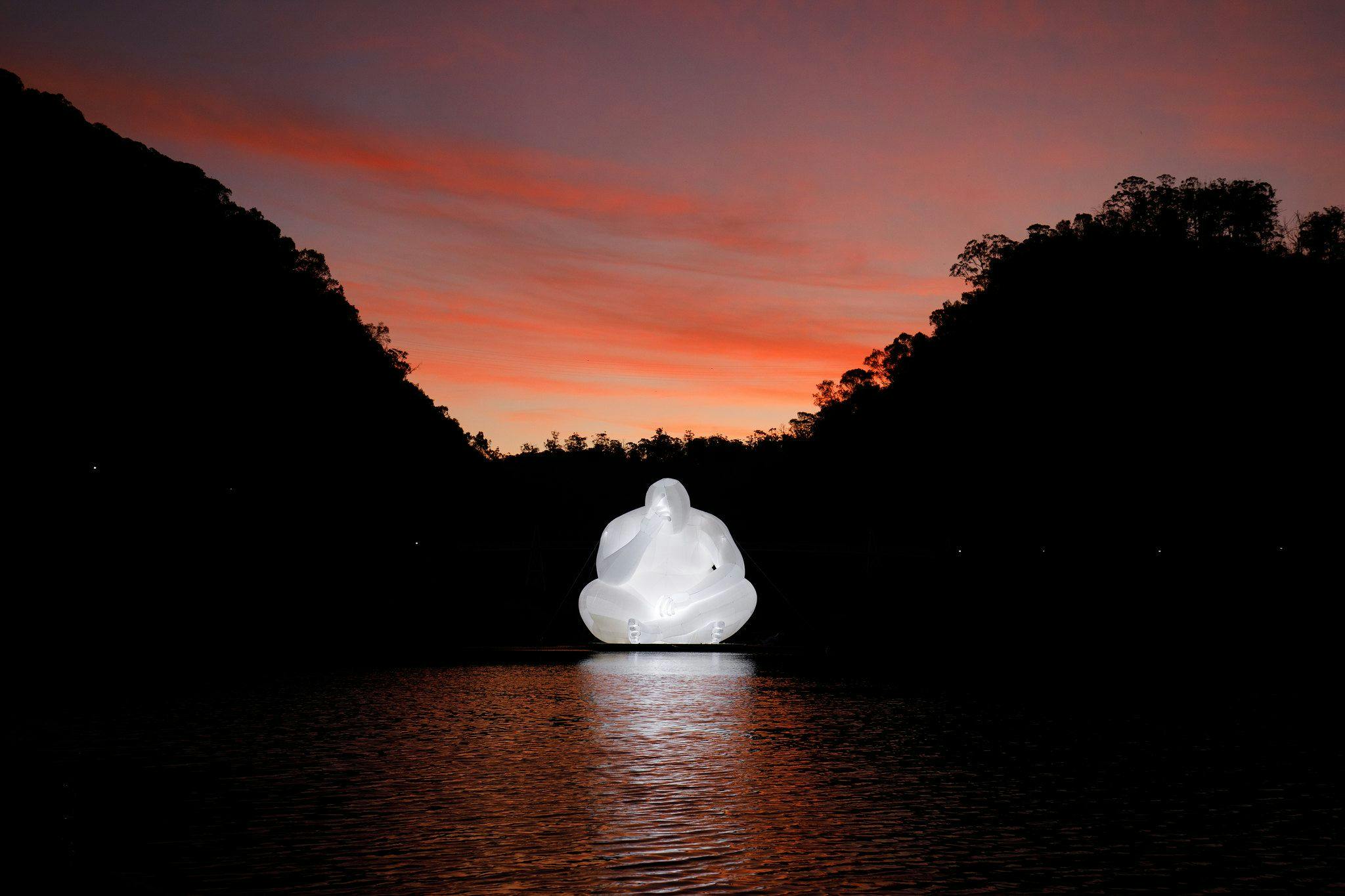 Sculpture of inflatable person head in hand on water at sunset.