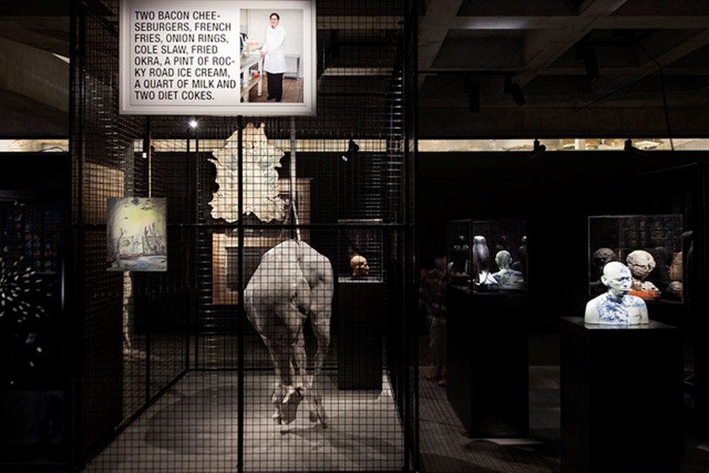 An elongated wax figure of a horse suspended from the ceiling of a black wire cage.