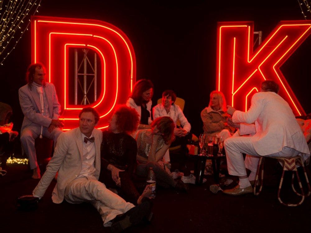 Wedding guests sitting around some tables having drinks. A large red neon D and K are lit up behind them.