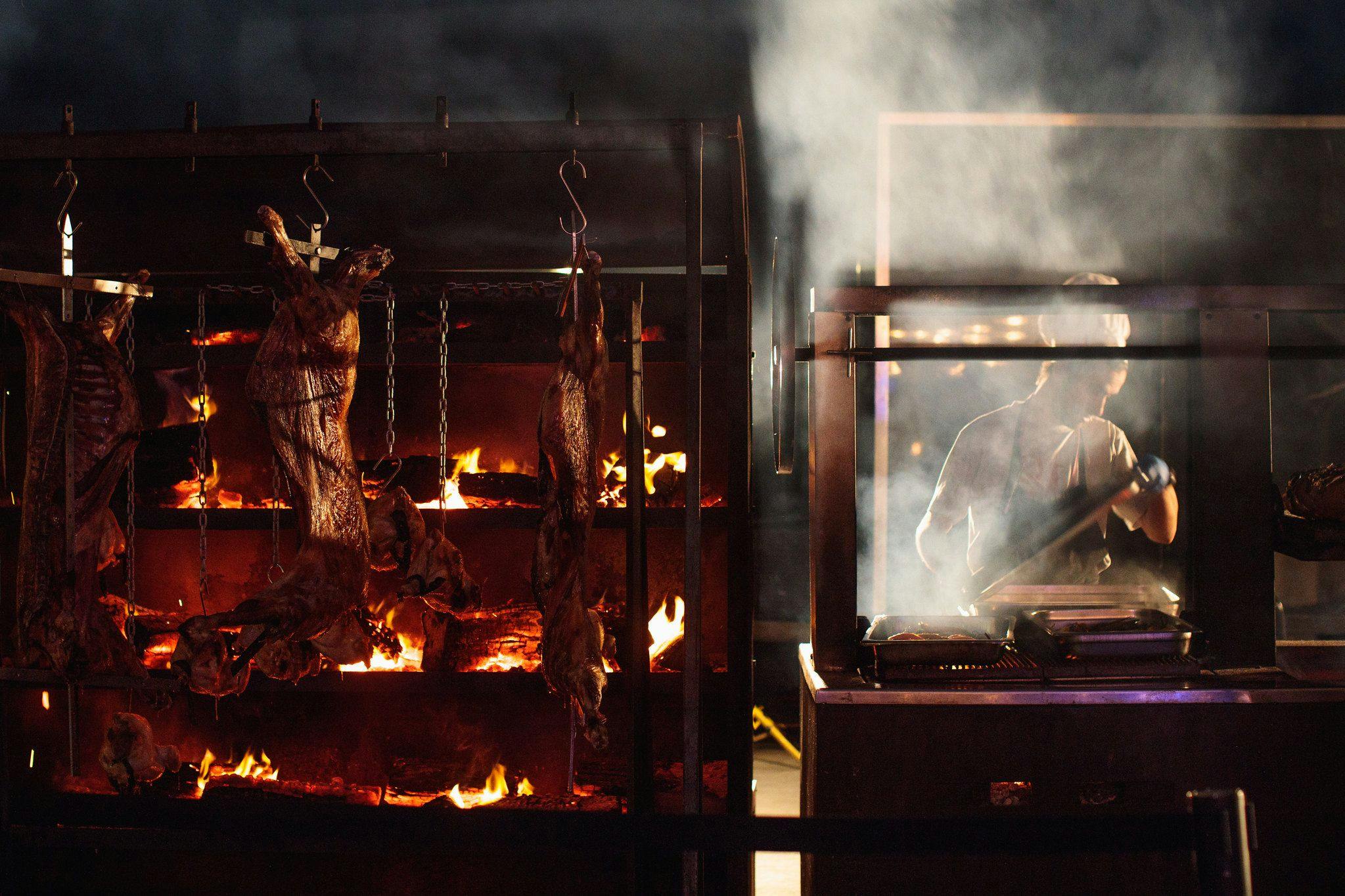 A tiered flaming grill  with meat being slowly cooked to perfection. A Chef standing to the right of the grill.