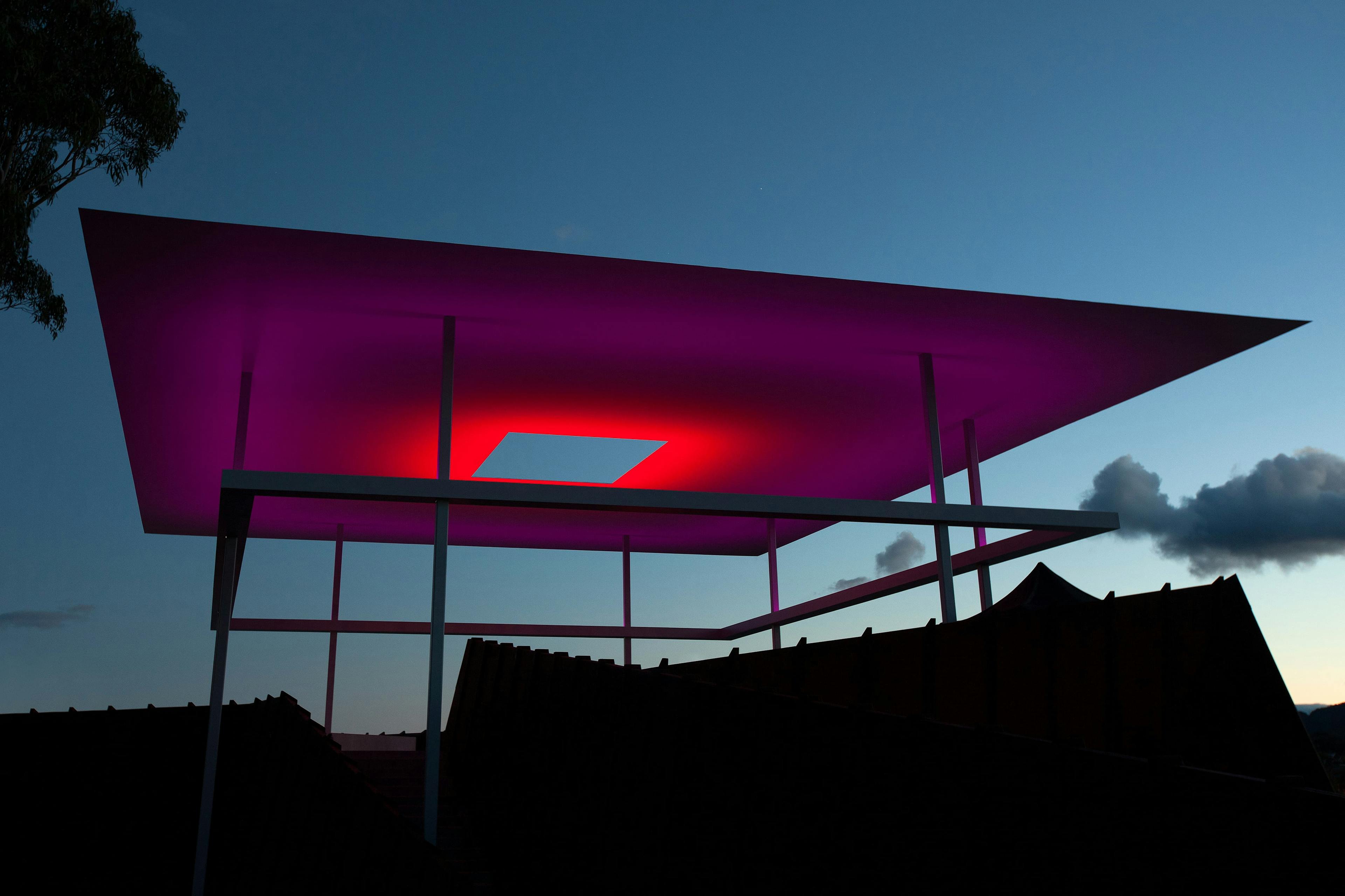 A twilight sky with a structure lit in pink and red with a hole in the centre.