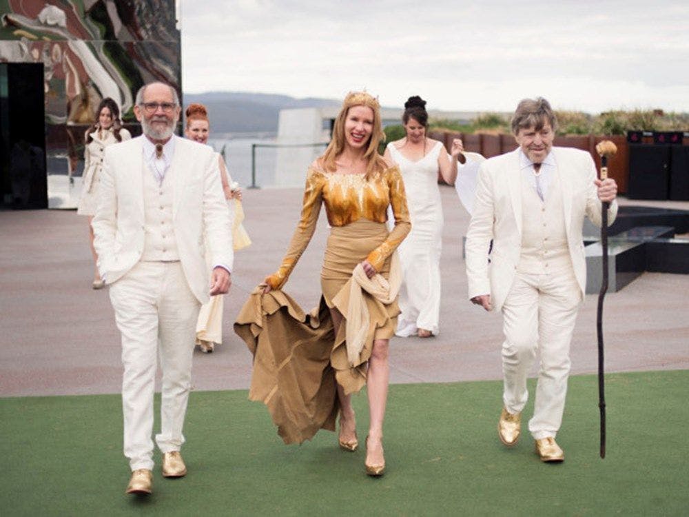 Kirsha standing in a golden wedding dress between two men in white suits.