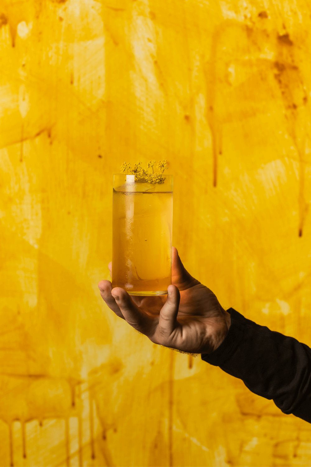A hand holding a high ball cocktail in front of a yellow wall
