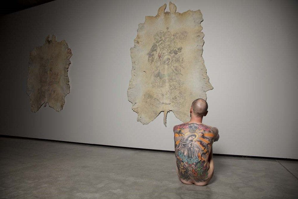 Man with tattooed back seated naked on concrete in front of an also tattooed pig skin.