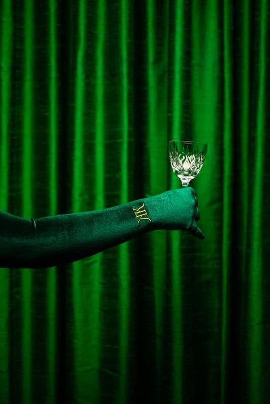 A green velvet gloved hand holding a crystal wine glass in front of green velvet curtains