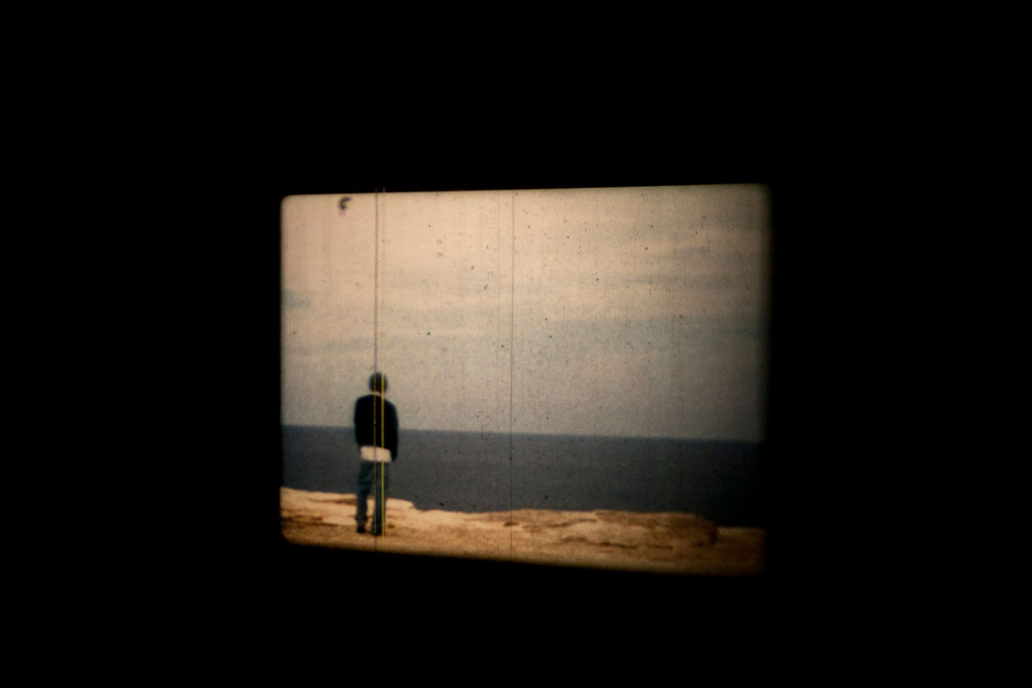 Still from a film. A person stands on sandstone rocks looking out to the ocean.