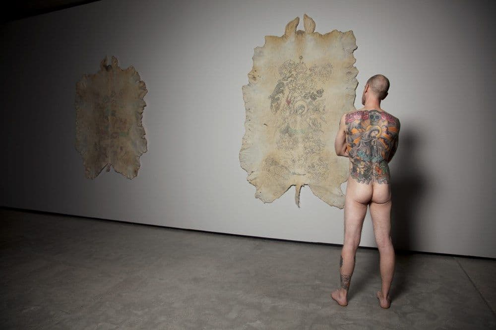 Tattooed naked man, back facing, standing in front of two hanging tattooed pigskins.