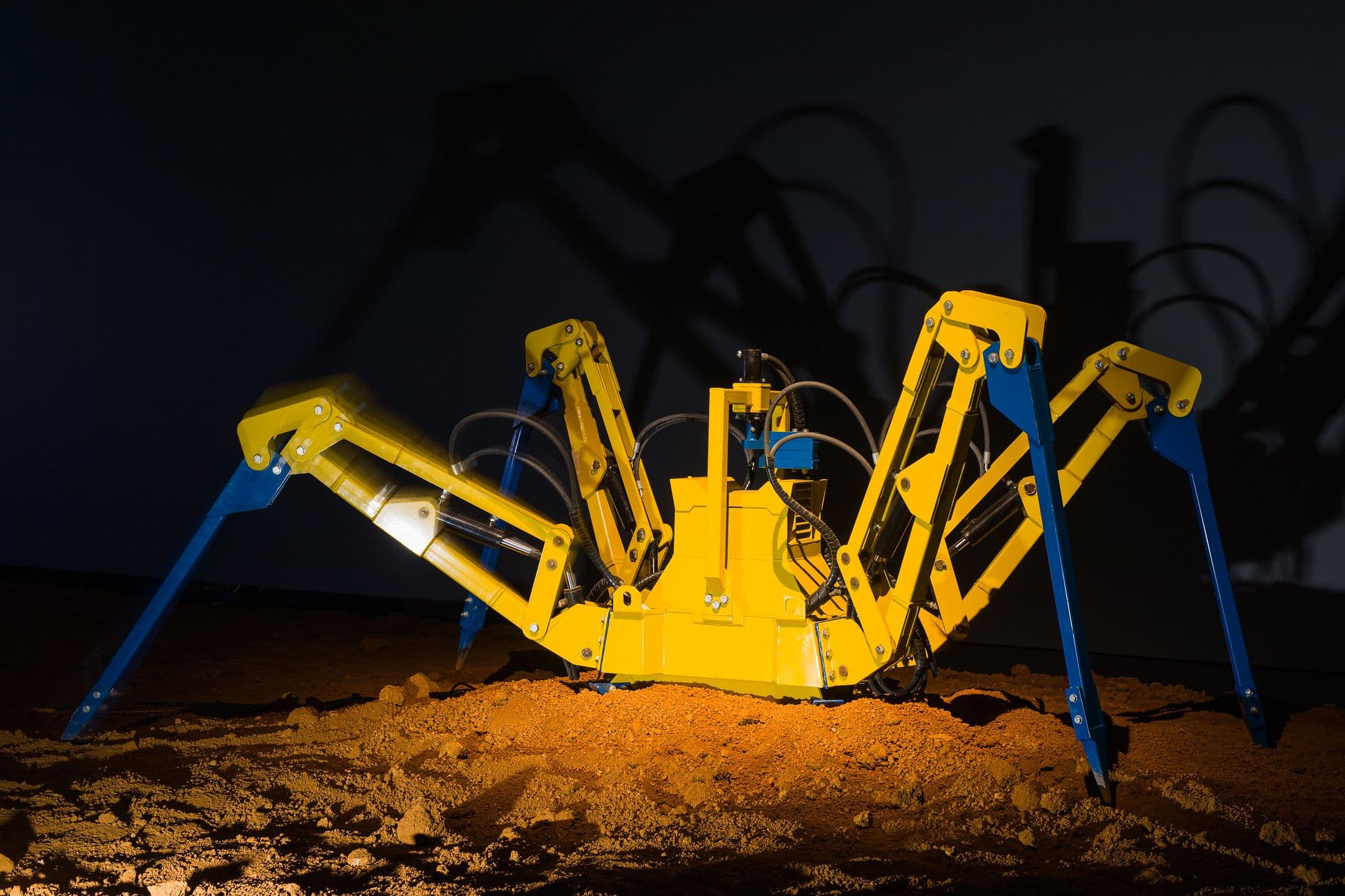 A yellow and blue hydraulic sculpture with 4 legs ontop of red dirt.