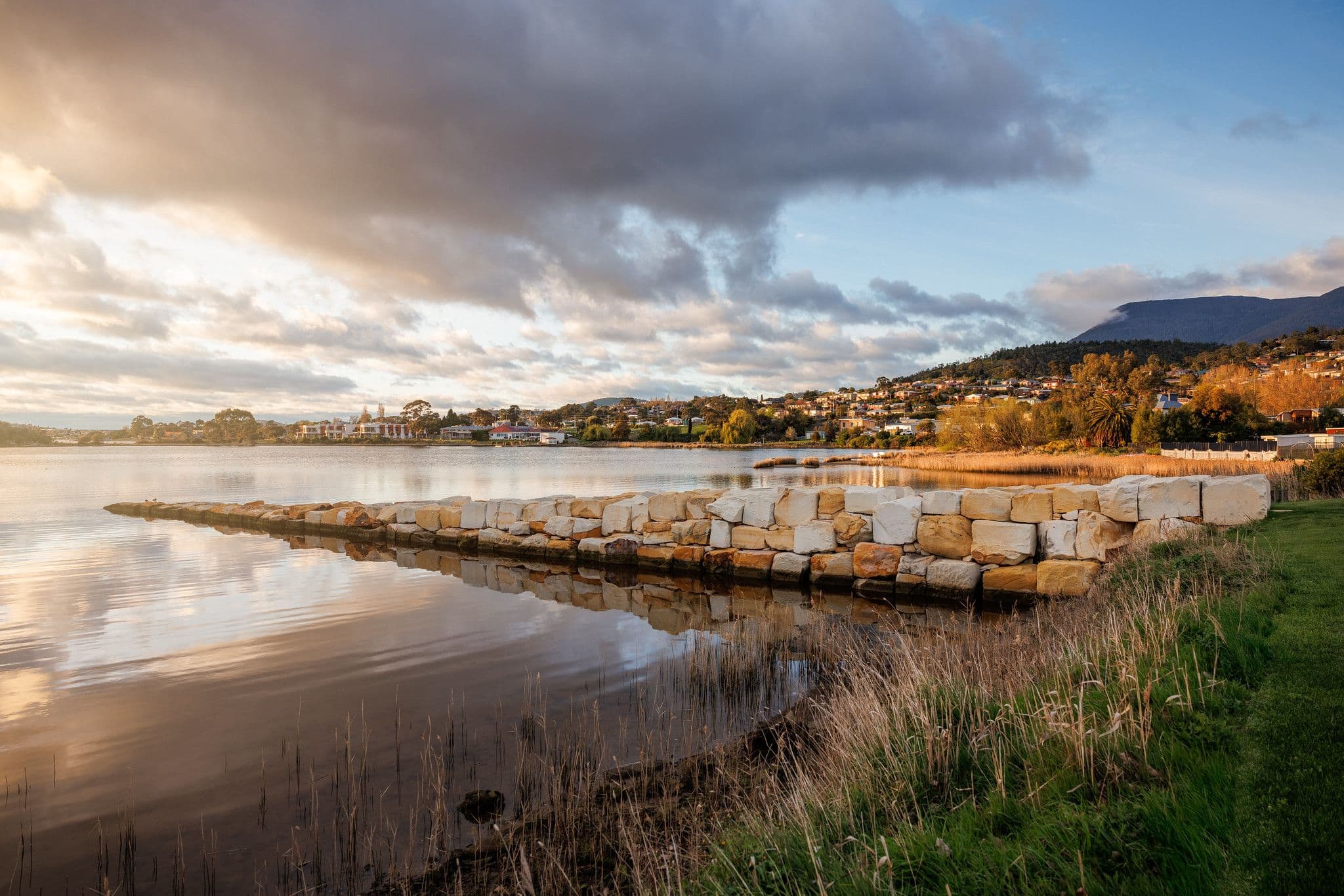 A stone path stretches from the shore into the Derwent River, in Berriedale, Hobart, Tasmania, with the backdrop of a sunrise.