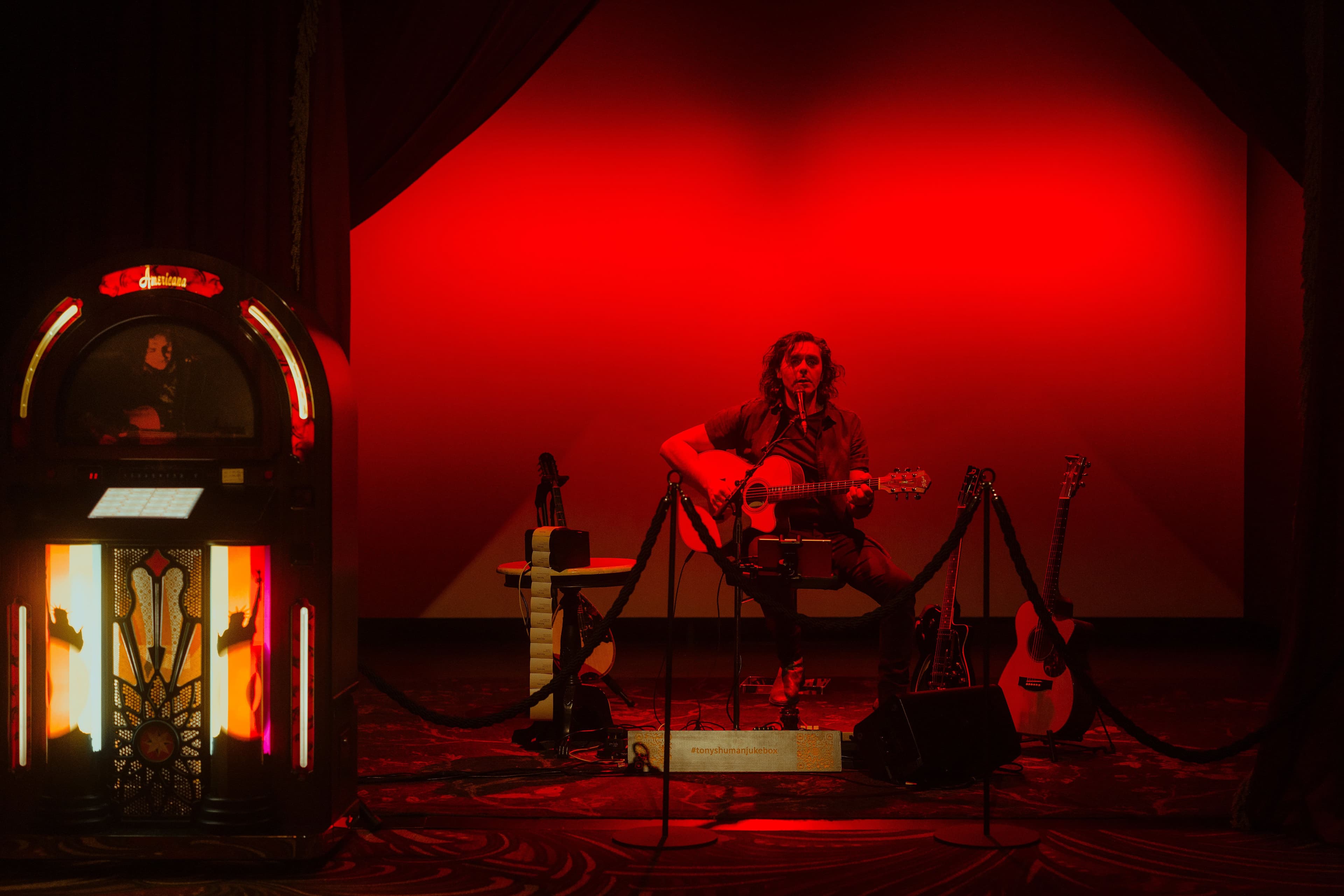 A guitarist plays, sitting on a stage in a red lit room. To his right, a lit-up vintage jukebox.