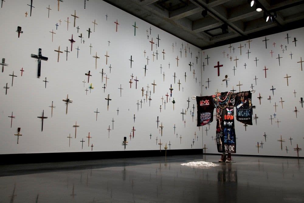 A black robe adorned with ACDC logos hands in a white room with crucifixes of all sizes and colours on the wall.