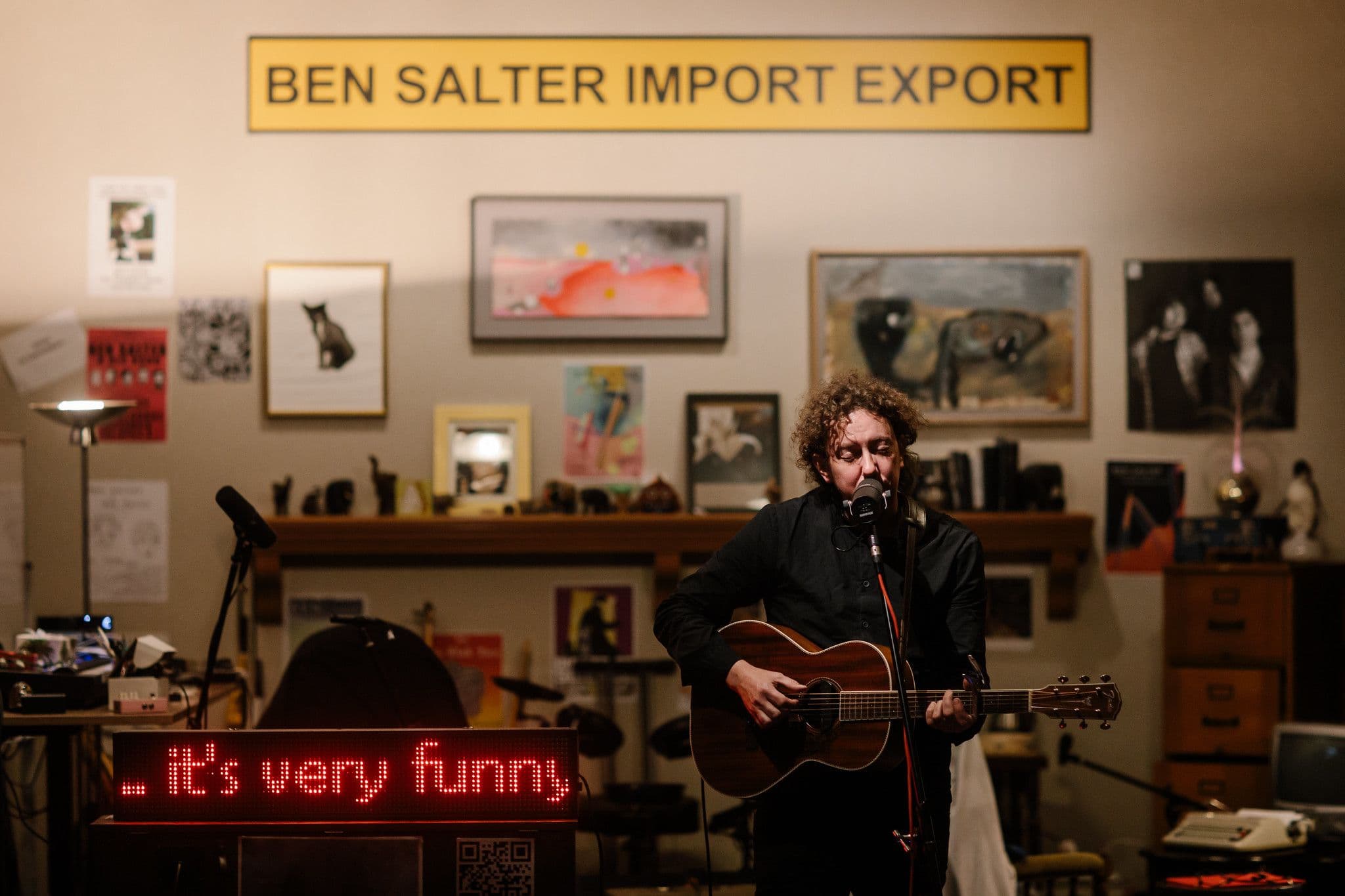 Ben Salter sings and plays guitar under a sign reading "BEN SALTER IMPORT EXPORT" and a screen reading "it's very funny"